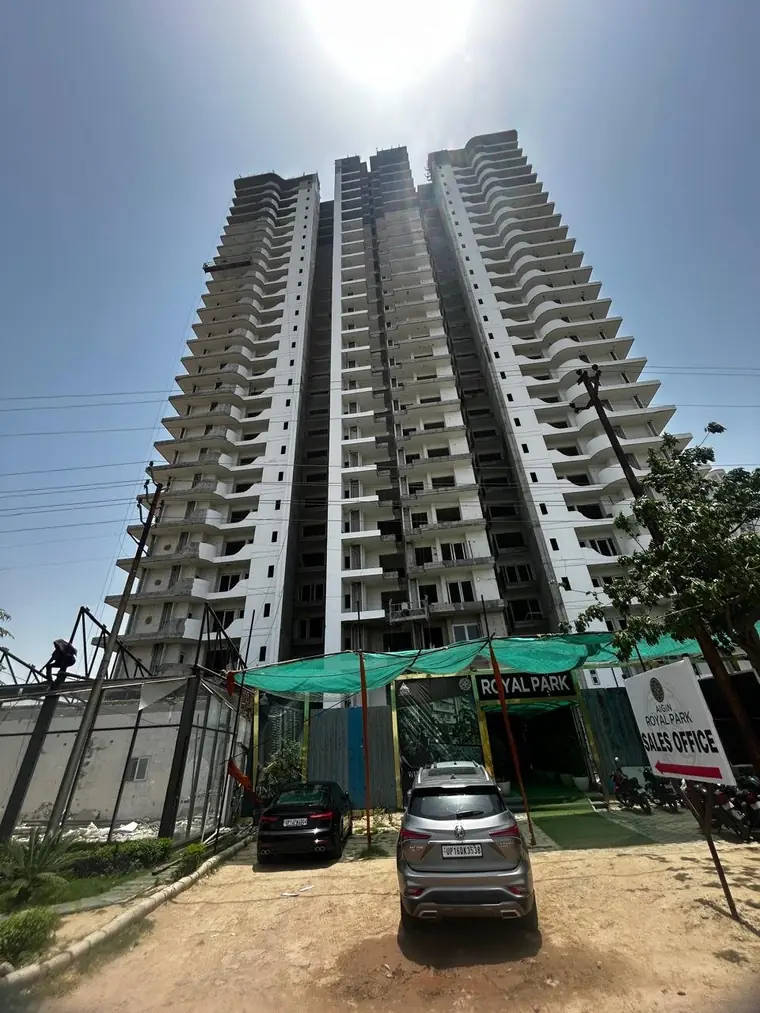 Both AIGIN Royal Park towers under construction at Aditya World City, NH-24 Ghaziabad, seen from the site entrance with the sales office canopy and visitor parking at their base
