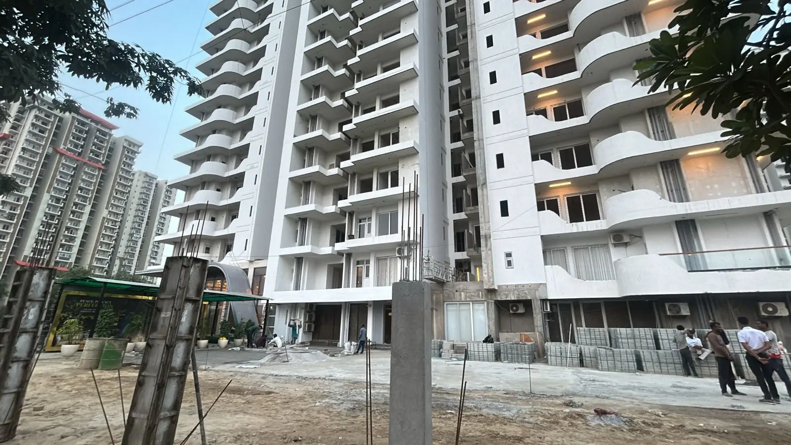 Entrance and lower floors of AIGIN Royal Park, Ghaziabad, photographed on site: white curved balconies above the entrance with glazing installed, and external works under way in the forecourt