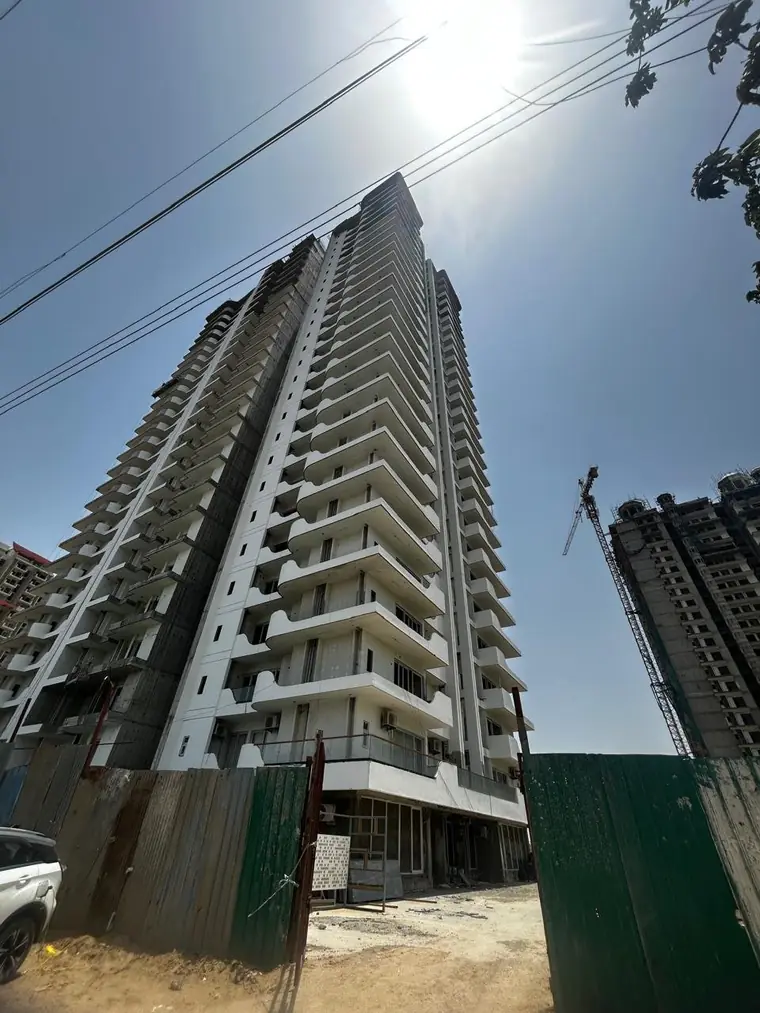 AIGIN Royal Park tower photographed from ground level against a clear sky, showing the completed G+27 structure with recessed balconies down the elevation and a second tower under crane behind