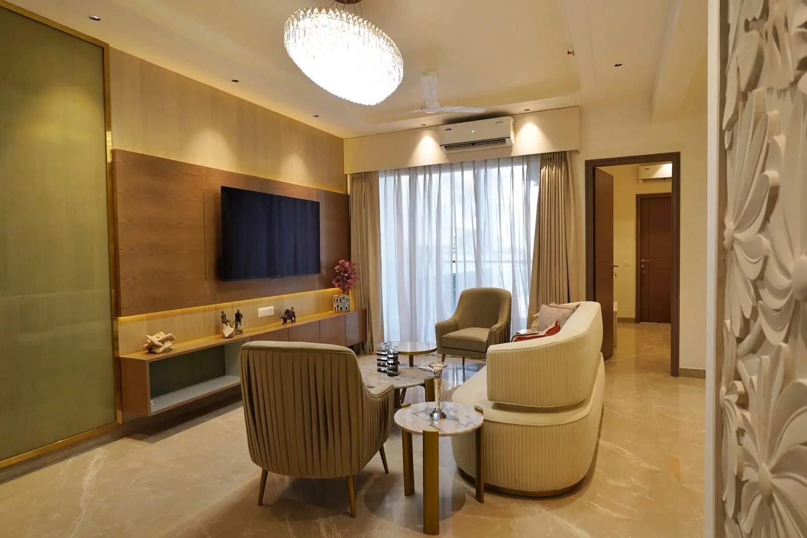 Living room in the finished sample flat at AIGIN Royal Park, Ghaziabad, with a television wall, chandelier and seating by the balcony